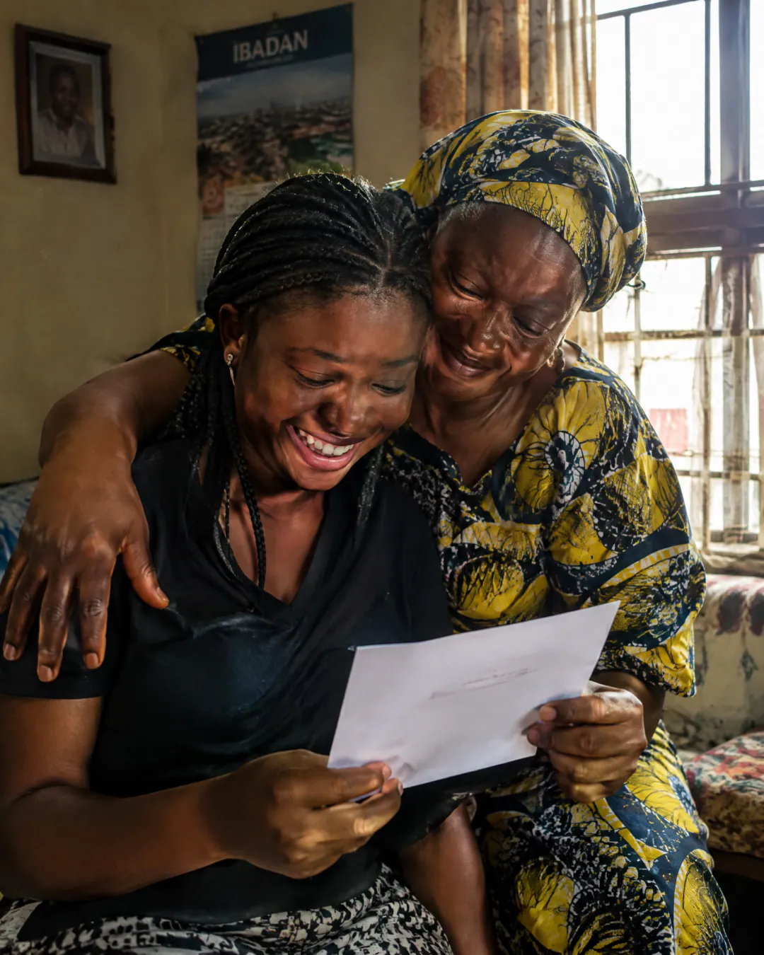 A laughing young woman reads a letter as an older woman in a yellow and navy headtie hugs her close at home.